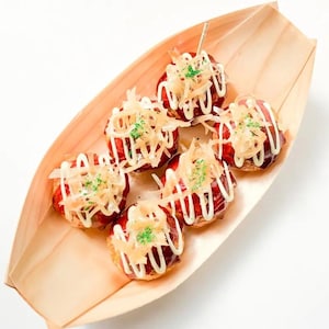 May include: Six takoyaki balls in a wooden boat-shaped dish. The takoyaki are topped with white sauce, shredded dried bonito flakes, and green garnish. A toothpick is inserted into one of the balls.
