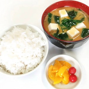 May include: A white bowl filled with white rice, a red and black bowl with a light brown broth, tofu, and green leafy vegetables, and a small white bowl with yellow pickled vegetables and red pickled fruit.