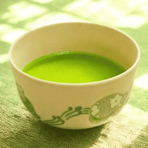 Może przedstawiać: Ceramiczna miska matcha wypełniona żywozieloną herbatą. Kremowa miska ma zielony kwiatowy wzór na obrzeżu. Herbata ma gładką, pienistą konsystencję, co wskazuje na prawidłowe ubicie.