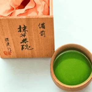 Puede incluir: Una caja de madera con caracteres japoneses y un pequeño sello rojo. Un pequeño cuenco redondo marrón lleno de té matcha verde brillante está junto a la caja. La caja contiene una tela color melocotón.