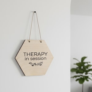 以下が含まれることがあります： 黒字で「THERAPY in session」と書かれた、明るい色の六角形の木製サイン。テキストの下には小さな花の模様があります。サインは白い壁に、細くて明るい色の紐で吊るされています。