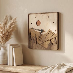 Handmade square plywood wall clock with layered mountain landscape, forest trees, sun detail, dark wood frame, black hands, and minimalist dot hour markers. Styled in a warm boho Japandi interior with beige wall, rustic wooden table, ceramic vase, pampas grass, neutral books, and linen textiles. Wooden mountain clock for cozy home decor.