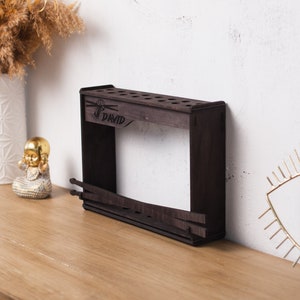 Pode incluir: Um suporte de baquetas de madeira castanha escura com o nome "DAVID" gravado na parte superior. O suporte tem vários orifícios para baquetas e uma secção inferior para armazenamento adicional. É exibido numa superfície de madeira.