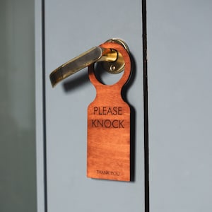 May include: A wooden door hanger with the text "Please Knock" and "Thank You" in a brown font. The hanger is hanging on a door handle.