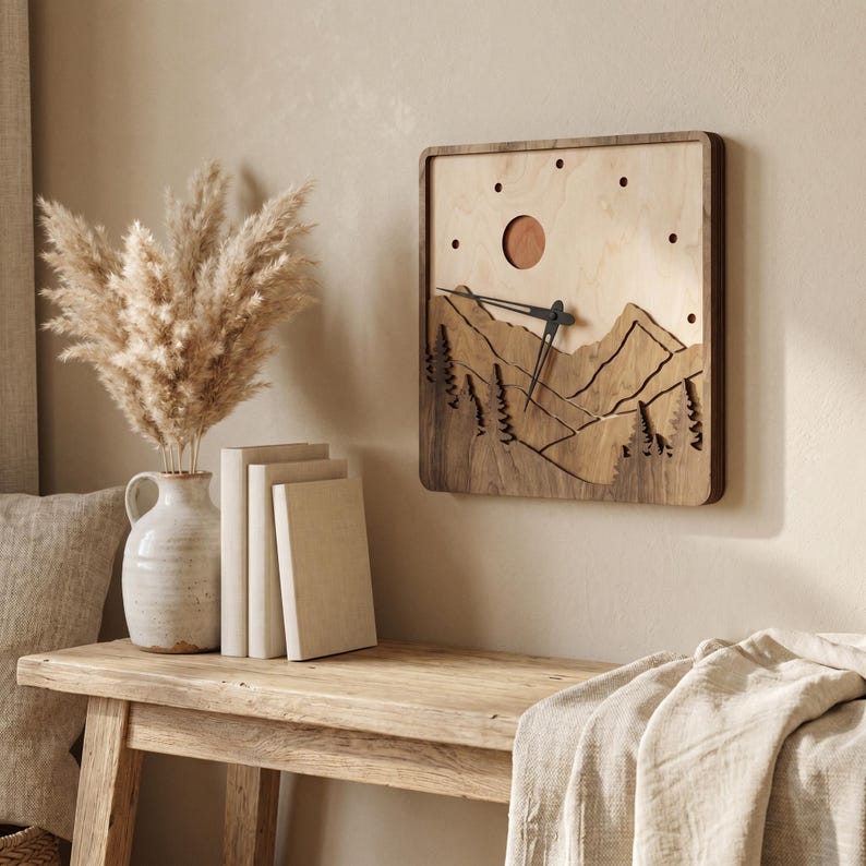 Handmade square plywood wall clock with layered mountain landscape, forest trees, sun detail, dark wood frame, black hands, and minimalist dot hour markers. Styled in a warm boho Japandi interior with beige wall, rustic wooden table, ceramic vase, pampas grass, neutral books, and linen textiles. Wooden mountain clock for cozy home decor.