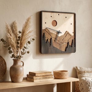 Handmade square plywood wall clock with layered mountain landscape, forest trees, sun detail, black hands, and minimalist dot hour markers. Styled in a warm boho Japandi interior with beige wall, wooden shelf, pampas grass, vintage books, woven basket, and soft sunlight. Rustic wooden mountain clock for cozy home decor.