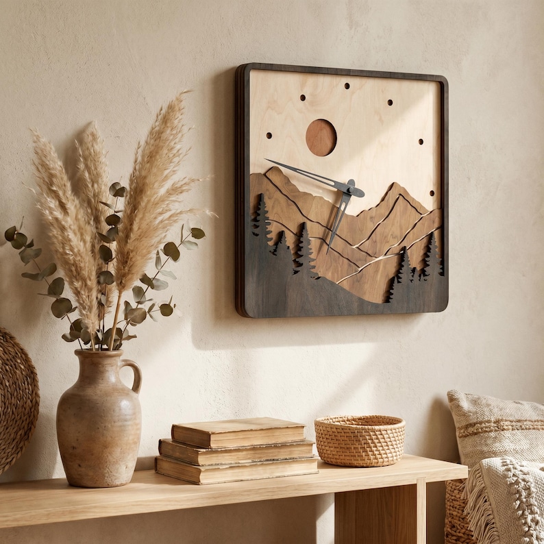 Handmade square plywood wall clock with layered mountain landscape, forest trees, sun detail, black hands, and minimalist dot hour markers. Styled in a warm boho Japandi interior with beige wall, wooden shelf, pampas grass, vintage books, woven basket, and soft sunlight. Rustic wooden mountain clock for cozy home decor.