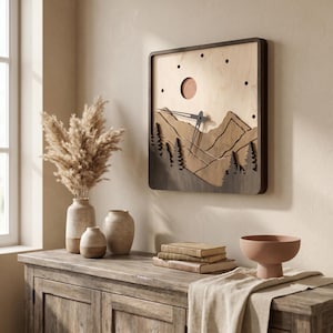 May include: A square wooden wall clock with a mountain scene. The clock features a dark brown frame, a light brown background, and a cut-out design of mountains and trees. The clock hands are black. The clock is mounted on a beige wall.