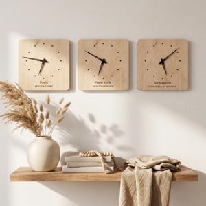 Puede incluir: Tres relojes cuadrados de madera con manecillas negras y marcadores de hora de puntos están montados en una pared blanca. Cada reloj muestra la hora de diferentes ciudades: París, Nueva York y Singapur. Una estantería de madera debajo contiene un jarrón, libros y objetos decorativos.