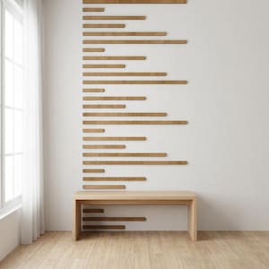 May include: A minimalist interior design featuring a light wood bench and a series of horizontal wooden planks mounted on a white wall. The planks vary in length and are arranged in a staggered pattern, creating a modern aesthetic. The room has a natural light from a window.