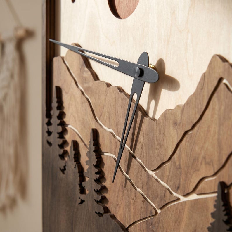 May include: Wooden wall clock with a mountain and forest design. The clock features a light wood background with dark wood cutouts of mountains and trees. The clock hands are black.