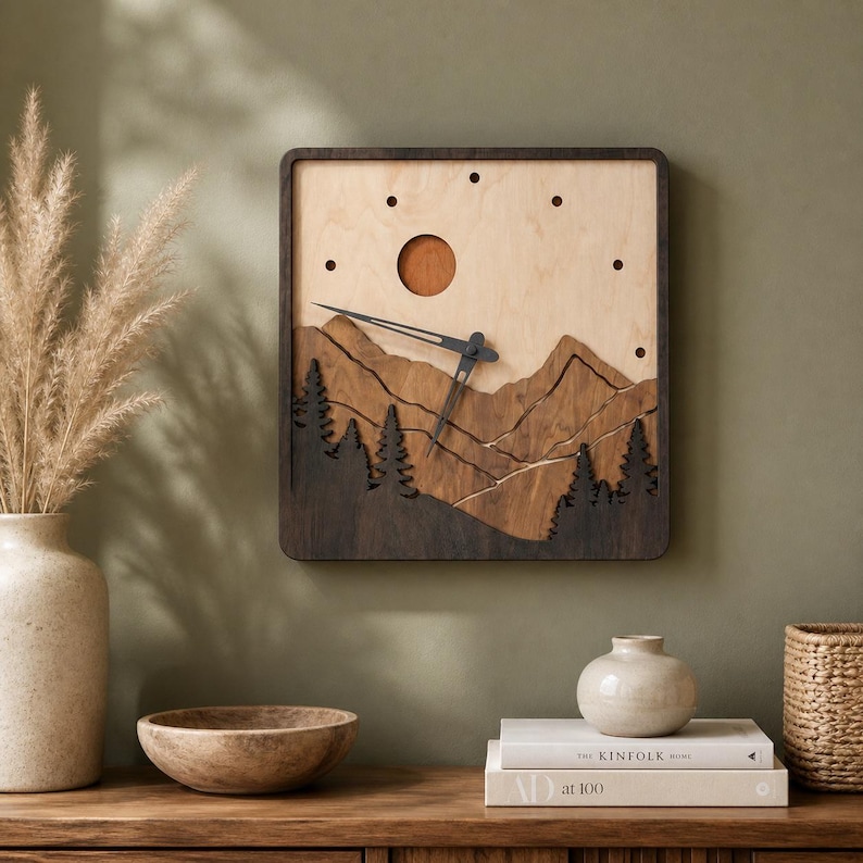 Handmade square plywood wall clock with layered mountain landscape, forest trees, sun detail, dark wood frame, black hands, and minimalist dot hour markers. Styled on a sage green wall in a warm boho Japandi interior with ceramic vases, pampas grass, books, woven basket, wooden decor, and soft sunlight. Rustic wooden mountain clock for cozy home decor.