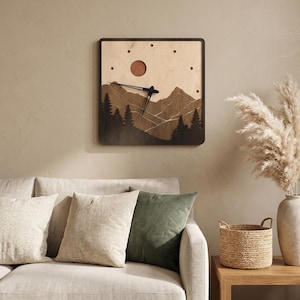 May include: Square wooden wall clock with a mountain landscape design. The clock features a dark brown frame, a light brown background, and a circular sun. The clock hands are black.