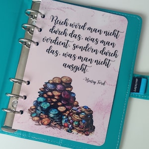 Könnte beinhalten: Eine rosa und weiße Planerseite mit einem Zitat von Henry Ford: "Reich wird man nicht durch das, was man verdient, sondern durch das, was man nicht ausgibt." Das Zitat ist von einem Haufen Goldmünzen mit Juwelen umgeben.