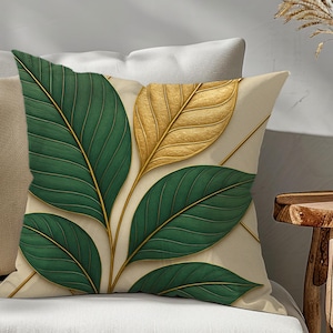 May include: A square throw pillow featuring a cream-colored background with a botanical print of large, dark green leaves and a single prominent gold leaf. The design includes thin, gold-colored stems connecting the leaves.