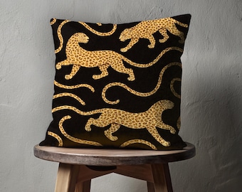 May include: A decorative square pillow with a black background and a repeating pattern of golden-colored leopards. The leopards are depicted in a running pose with their tails curved. The pillow is placed on a small wooden stool.