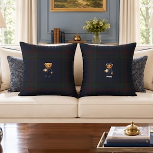 May include: Two square throw pillows featuring a dark navy blue, green, and red plaid pattern. Each pillow displays a centered embroidered teddy bear graphic. The left bear wears a navy coat, while the right bear wears a navy dress with a bow.