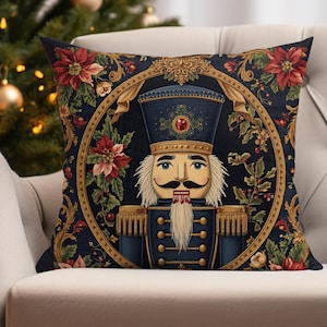 May include: Decorative square pillow featuring a detailed illustration of a nutcracker soldier in a blue uniform and hat. The design is framed by a gold border and surrounded by red poinsettias and green foliage, creating a festive holiday theme.