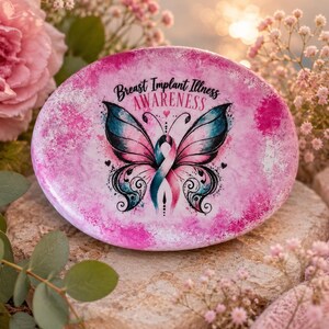 May include: A pink, oval-shaped stone with the words "Breast Implant Illness Awareness" and a butterfly design. The butterfly wings are pink and teal, with a pink and teal ribbon in the center.