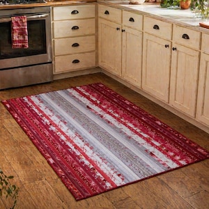 May include: A rectangular rug with a striped pattern in shades of red, white, and gray. The rug is placed on a wooden floor in a kitchen setting. The rug's design features a series of parallel lines.