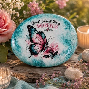 May include: A decorative, oval-shaped stone with a teal and white background. The stone features a butterfly with pink and blue wings and the words "Breast Implant Illness Awareness". The stone is surrounded by flowers and candles.