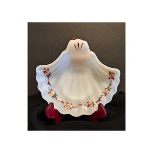 May include: White milk glass shell-shaped dish with a brown and pink floral design around the rim. The dish is on a wooden stand.