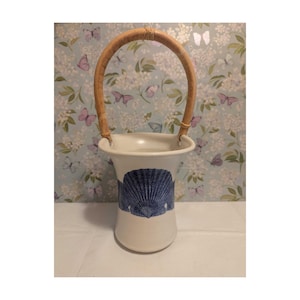 May include: A white ceramic basket with a flared rim and a curved, light brown woven handle. The exterior of the vessel features a dark blue graphic print of a large scallop shell centered on the front, providing a clear contrast to the white base.