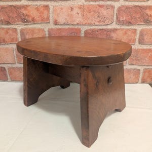 May include: A small, low-profile wooden stool with an oval-shaped seat and four sturdy, slightly flared legs. The stool features a dark brown, natural wood grain finish with visible joinery details, including round pegs on the side supports.