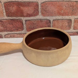 May include: A beige ceramic soup bowl with a handle. The interior is a dark brown color. The bowl is sitting on a white surface in front of a brick wall.