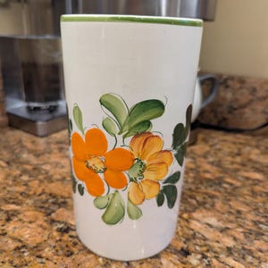 May include: A white ceramic tumbler with a green rim and floral design. The tumbler features orange and yellow flowers with green leaves. The tumbler is cylindrical and appears to be hand-painted.