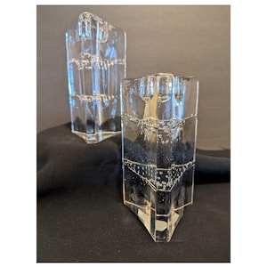 May include: Two clear glass candle holders with a geometric design. The candle holders are rectangular with a textured surface and a square top. The candle holders are on a black surface.