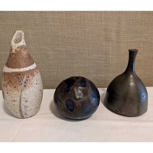 May include: Three ceramic vases of varying shapes and colors. The tallest vase is a speckled brown and white with a curved opening. A round, dark blue vase sits next to it, and a dark gray vase with a narrow neck is on the right.