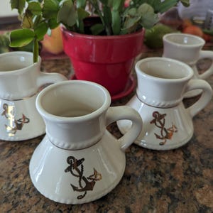 May include: A set of four off-white ceramic mugs with a wide, flared base and a single handle. Each mug features a metallic gold anchor graphic on the front and a subtle rope-like texture detail around the middle section of the exterior body.