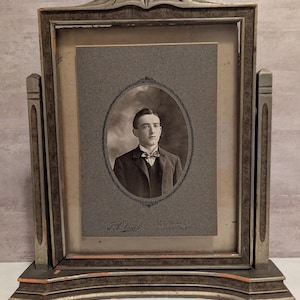 May include: An antique portrait frame with a black and white photograph of a man in a suit and bow tie. The frame is made of wood with a distressed finish in shades of gray and brown. The photograph is in an oval frame.