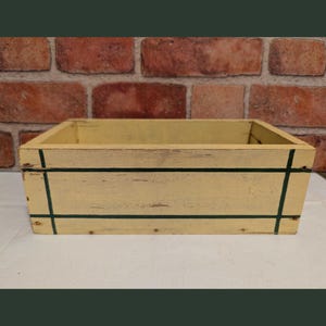 May include: A rectangular wooden crate with a distressed cream-colored finish. The box features thin, dark green painted lines forming a grid pattern across the front and sides. The wood grain is visible beneath the paint, showing signs of wear.