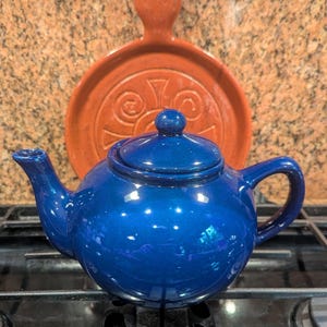 May include: A glossy, deep blue ceramic teapot with a matching lid, featuring a curved handle and a short spout. Positioned directly behind the teapot is a round, terracotta-colored ceramic plate with a central embossed spiral design.