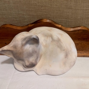 May include: A ceramic cat figurine in a curled-up position, painted in shades of cream and gray. The cat is resting on a white surface, with a wooden shelf in the background. The figurine has a smooth, matte finish.