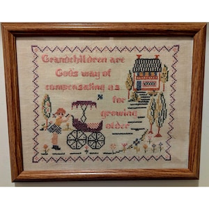 May include: Framed cross-stitch artwork with a brown wooden frame. The artwork features a house, trees, a child with a stroller, and the text "Grandchildren are God's way of compensating us for growing older."
