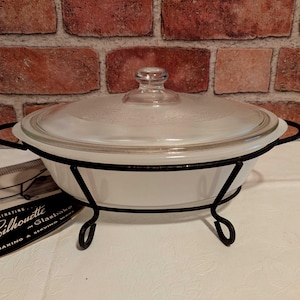 May include: A white oval Glasbake casserole dish with a clear glass lid, resting in a black metal wire stand with curved handles and feet. Beside the dish is a small black and white informational card labeled Fascinating Silhouette in Glasbake.