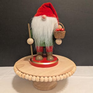May include: A decorative gnome figurine with a red velvet hat, white beard, and green tunic. The gnome holds a small basket with red mushrooms and a wooden staff. It stands on a wooden pedestal with a beaded edge, perfect for holiday decor.
