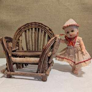 May include: A small doll with blue eyes and a light complexion wearing a cream-colored dress with red polka dots and a matching bonnet, standing next to a miniature chair constructed from brown twigs and branches with a curved backrest design.