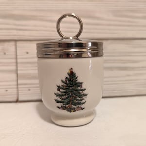 May include: A white ceramic egg cup with a silver-colored lid and handle. The egg cup features a Christmas tree design with ornaments and presents. The egg cup is a festive holiday decoration.