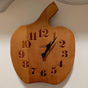 May include: A light brown wooden wall clock shaped like an apple with a stem at the top. The clock face features dark brown cutout numerals from one to twelve and the word Linder printed in the center. Two black hands indicate the time.