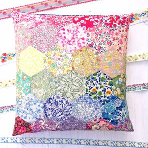 May include: A square throw pillow constructed from a patchwork of hexagonal fabric pieces. Each hexagon features a distinct floral pattern in a variety of colors including pink, yellow, green, blue, and purple. The pillow is surrounded by ribbons.