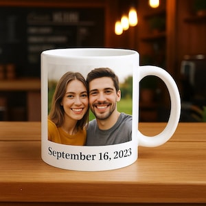 Può includere: Tazza in ceramica bianca con una foto di una coppia sorridente e la data "16 settembre 2023". La tazza ha un manico curvo ed è appoggiata su una superficie di legno.