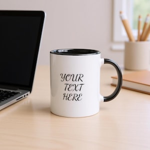 Può includere: Tazza in ceramica bianca con interno e manico neri. La tazza presenta il testo "YOUR TEXT HERE" in corsivo nero. La tazza è su una superficie di legno chiaro, accanto a un laptop e ad altri articoli per ufficio.
