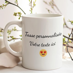 May include: White ceramic mug with a curved handle. The mug features the text "Tasse personnalis&eacute;e Votre texte ici" in black script, along with a yellow emoji with heart eyes. The mug is set against a blurred background with greenery.