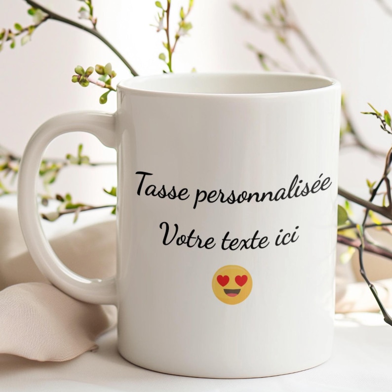 May include: White ceramic mug with a curved handle. The mug features the text "Tasse personnalis&eacute;e Votre texte ici" in black script, along with a yellow emoji with heart eyes. The mug is set against a blurred background with greenery.