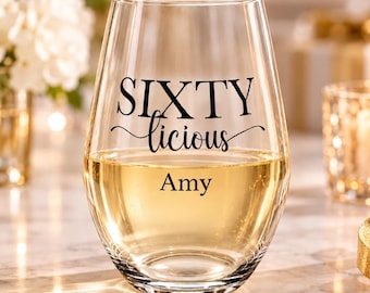 May include: A clear stemless wine glass featuring the black printed text SIXTY licious and the name Amy below it. The glass is partially filled with a light-coloured liquid, showcasing a smooth, rounded silhouette and a transparent glass construction.