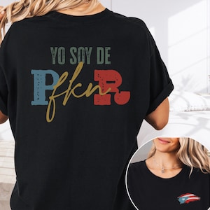 Peut inclure: T-shirt noir avec l'inscription "YO SOY DE PR" dans un style vieilli. Les lettres "P" et "R" sont bleues et rouges, avec un script doré "fkn" entre les deux. Un petit drapeau portoricain est sur le devant.